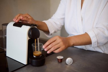 Beyaz waffle bornozlu genç bir kadının sabah espresso 'sunu ev kullanımı için kapsül kahve makinesiyle yaparken yakın çekimi. Güne enerji veren kafein içeceğiyle başlarsın. - İnsanlar. Yiyecek ve içecek.