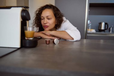 Güzel, kıvırcık saçlı esmer, beyaz bornozlu genç, yetişkin, güzel bir kadın mutfak tezgahına yaslanıyor ve kapsüllü kahve makinesine hayran, mutfakta kahvaltı için kahve yapıyor.