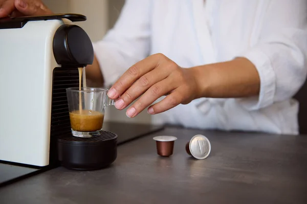 Close-up of a housewife\'s hands using a capsule coffee machine preparing freshly brewed espresso coffee, standing at the kitchen counter in cozy home. Start your day with an energizing cup of coffee.