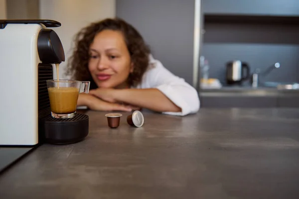 Details on a transparent glass cup with freshly brewed espresso coffee with foam and happy woman enjoying making energizing hot drink, using capsule coffee machine for home use. Morning routine