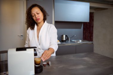 Kıvırcık, koyu renk saçlı, çok ırklı, uykulu, beyaz bornozlu, sabahları kahve makinesiyle taze demlenmiş kahve hazırlayan genç bir kadın. Sabah rutini. - İnsanlar. Yaşam tarzları. Boş zaman