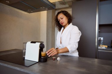 Beyaz bornozlu, kapsül kahve makinesi kullanan çok ırklı, uykulu bir kadının portresi sabah espresso 'sunu mutfağında yapıyor.