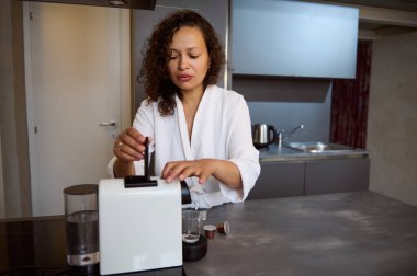 Güzel, uykucu genç bir kadın kahve makinesinin içine kahve koyarak sabah kahvaltısı için taze demlenmiş espresso hazırlıyor, mutfak tezgahında duruyor.