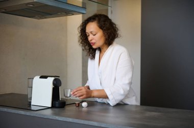 Beyaz bornozlu genç bir kadının özgün portresi kahvaltıda kapsül kahve makinesi kullanarak espresso kahvesi yapıyor. - İnsanlar. Boş zaman. Yaşam tarzı. Güne enerji dolu bir kahveyle başla.