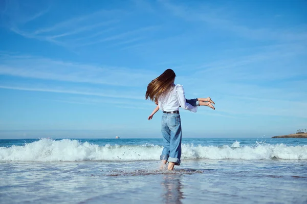Full length portrait of a mother with long hair, in blue denim, playing with her little child daughter, spinning her around, running barefoot on the waves on Atlantic ocean beach. Happy family pastime