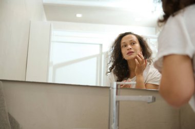 Genç, çok ırklı, sağlıklı, ışıl ışıl cilde sahip bir kadın banyo aynasında yansımasına bakarken sabah spa prosedürleri sırasında yüzüne nemlendirici köpük sürüyor.