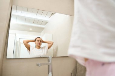 Genç ve güzel bir kadın aynada yansımasına bakarken nemlendirici ve temizleyici köpük sürüp yüzüne parlak, temiz bir ciltle kozmetik sürüyor. Sabah rutini, güzellik ve spa