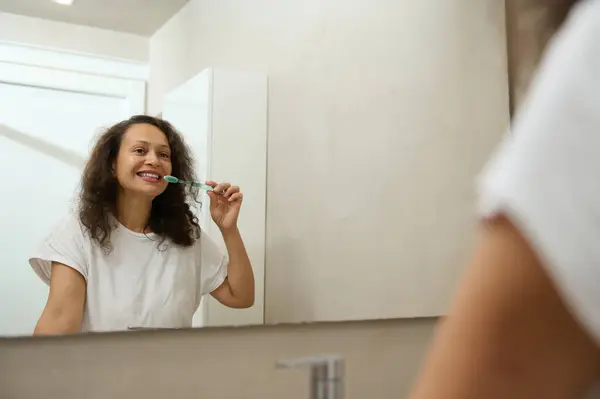 Reflection in the mirror of a happy confident good looking young adult woman in white pajama, brushing teeth and smiling, admiring her appearance. Healthcare and medicine. Dental care and oral hygiene