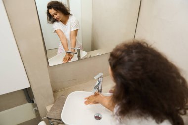 Aynada yansıyan güzel bir kadın, akan suyun altında antibakteriyel sabun köpüğüyle ellerini yıkıyor, ev banyosundaki seramik beyaz lavaboda kurnazca gülümsüyor. Deri, vücut bakımı ve hijyen.