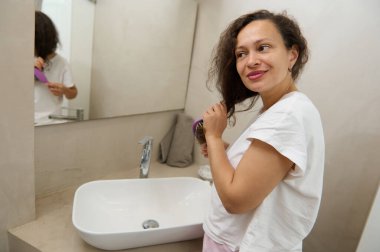Beyaz tişörtlü çok ırklı güzel bir kadın banyoda aynaya bakarken saçlarını tarıyor ve gülümsüyor. Saç, güzellik ve kozmetik. Tedavi ve bakım.