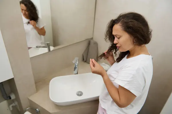 Young adult woman adjusting and examining her damaged hair in bathroom mirror in morning. Worried young woman reflected in home mirror, concerned about hair loss. Haircare, health problem concept