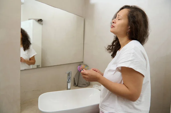Worried young woman reflected in home mirror, concerned about hair loss, combing at mirror, holding hair brush and lock. Upset young woman counting fell out hairs. Haircare, health problem concept