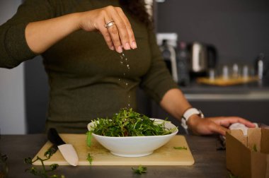 Ev mutfağında öğle yemeği hazırlarken kadın elinin tuz tutmasına ve taze sağlıklı salata baharatına odaklan. Yemek pişiren kadın. Arugula yapraklı beyaz bir seramik tabak ve olgun organik domatesler.