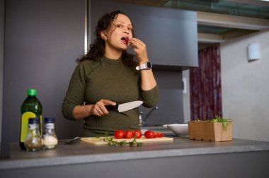 Çok etnik, genç ve güzel bir kadın sebze doğrarken kırmızı organik domatesleri tadıyor, mutfak adasında duruyor ve öğle yemeği için çiğ vegan salatası hazırlıyor. Sağlıklı beslenme, zayıflama ve diyet konsepti