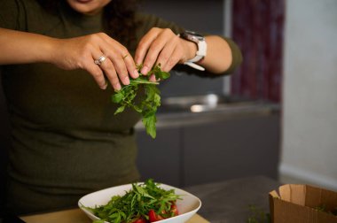 Ev mutfağında akşam yemeği pişirirken taze sulu domateslerle dolu bir kaseye roka yaprakları ekleyen bir kadının yakın çekim elleri. - İnsanlar. Sağlıklı yiyecekler. Zayıflatma ve diyet konsepti. Ev hayatı.