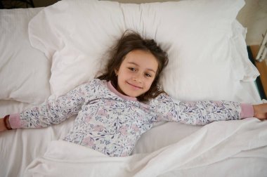 Otantik, mutlu, küçük bir kızın yatağında uzanmış kameraya bakıyor ve sabah uyandığında kollarını yukarı doğru açıyor.