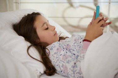 Yakın plan bir portre, beyaz bir ilkokul öğrencisi, akıllı cep telefonuyla eğlenen sevimli küçük bir kız sabah uyanırken yatakta yatıyor. Dijital cihaz bağımlılığı