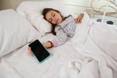 Pijamalı beyaz bir kız çocuğu elinde cep telefonuyla yatağında uyuyakalıyor. Siyah boş dijital dokunmatik ekranlı akıllı telefon. Dijital aygıt ve çevrimiçi video oyunları bağımlılığı