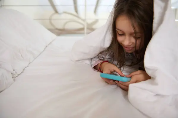 stock image Curious mischievous little kid girl in pajamas, playing online video games, washing cartoons, checking social media content on her smart mobile phone, hiding and lying down under white blanket.