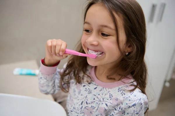 穿着睡衣的可爱的小女孩 拿着牙刷和牙膏 在家里浴室的镜子前刷牙 健康的生活方式牙科口腔护理及卫生 免版税图库图片