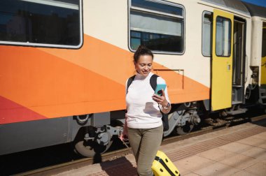 Çantası ve sırt çantasıyla gülümseyen turist kadın trene binerken akıllı telefondan mobil uygulamaları kontrol ediyor tren istasyonunda tren platformu boyunca yürüyor. İnsanlar ve gezi konsepti