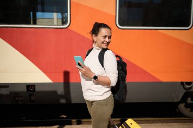 Tren istasyonunda tren beklerken cep telefonu kullanan gülümseyen genç Afrikalı Amerikalı kadın. - İnsanlar. Yaşam tarzları, seyahat konsepti. Tren ve demiryolu araçları ucuz taşımacılık gibi