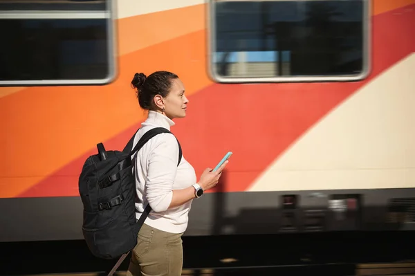 Side portrait multiracial woman commuter with suitcase and backpack, holding mobile phone, looking away, standing at railway platform while travelling. Train as cheap Railroad transport. Tourism