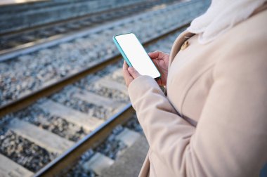 Beyaz, boş, dijital dokunmatik ekranlı modern bir akıllı telefonun detayları, fotokopi reklam alanı, bir kadın banliyöde tren istasyonunun platformunda tren bekliyor.
