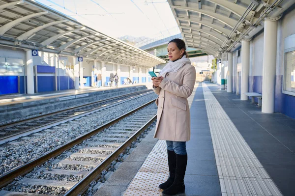 Full length portrait of a confident young woman in warm winter clothes, checking mobile app on her smartphone, online booking tickets while standing outdoor on the train platform of a railway station