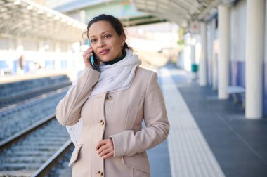 30 'lu yaşlardaki çok ırklı güzel bir kadın, tren istasyonunda tren beklerken cep telefonu kullanıyor, sohbet ediyor. Halk, teknolojiler ve toplu taşımacılık