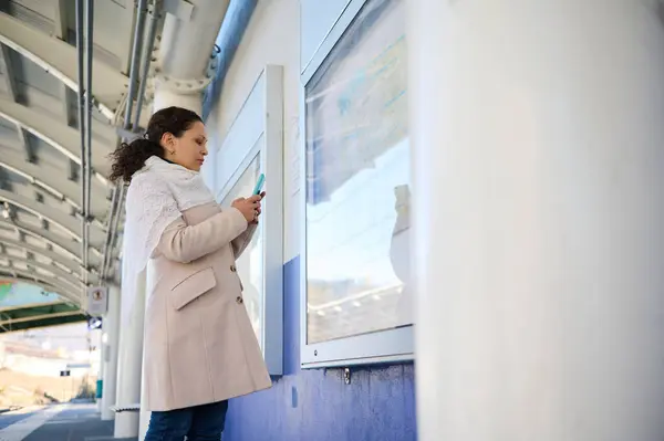 Multi ethnic young adult woman commuter using mobile phone, standing at information desk with train schedule on the platform of a railway station. People. Trip. Travel. Railroad transport concept