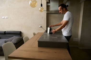 Beyaz, genç, yakışıklı adam sabah kahvesini evde kullanmak için kapsül kahve makinesiyle hazırlıyor. Mutfak tezgahında minimalist, rahat bir evin içinde duruyor.