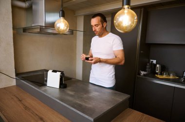 Beyaz tişörtlü çekici genç Latin Amerikalı adam, elinde cam bardak dolusu taze kahve, evinde mutfak masasında duruyor.