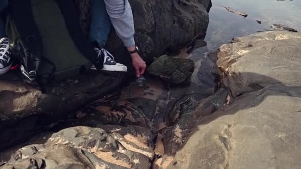 セラスフラスコのボトルからカップを持っている男性の冒険旅行者の手のクローズアップは 自然の中で岩の上に座って 海水でそれを洗って洗います 人々よ ピクニック ライフスタイル レクリエーション — ストック動画