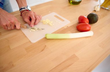 Erkek elleri kesme tahtasında sarımsak karanfili doğruyor, ahşap masada taze domatesler, avokado meyveleri ve pırasa ile duruyor, mutfakta sağlıklı diyet salata pişiriyor. Boşluğu kopyala