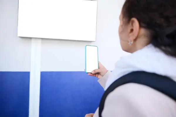 Focus on smartphone with white blank digital mockup screen in the hand of a woman commuter taking photo of the information desk with train schedules, standing on the platform of a railway station
