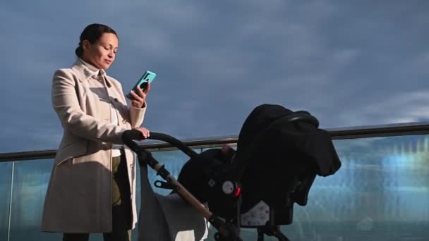 年轻漂亮的女人 妈妈推婴儿车 在户外用智能手机 — 图库视频影像
