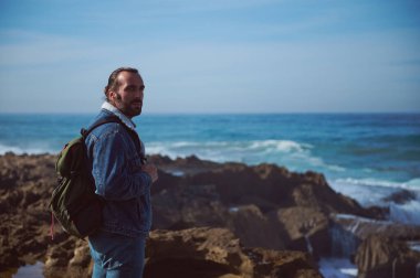 Sırt çantası taşıyan beyaz sakallı bir gezgin uçurumda dikilip kameraya bakıyor ve Atlantik Okyanusu 'nun güzel manzarasının tadını çıkarıyor. - İnsanlar. Turizm turizmi. Seyahat ve aktif yaşam tarzı kavramı