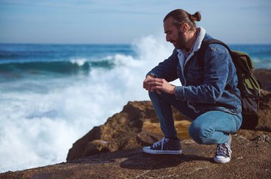 Yakışıklı genç turist kol saatini kontrol ediyor, dalgalarla deniz kenarındaki kayalıklarda dinleniyor. - İnsanlar. Seyahat et. Güzel vahşi doğayı keşfetmek. Aktif turizm ve yaşam tarzı konsepti