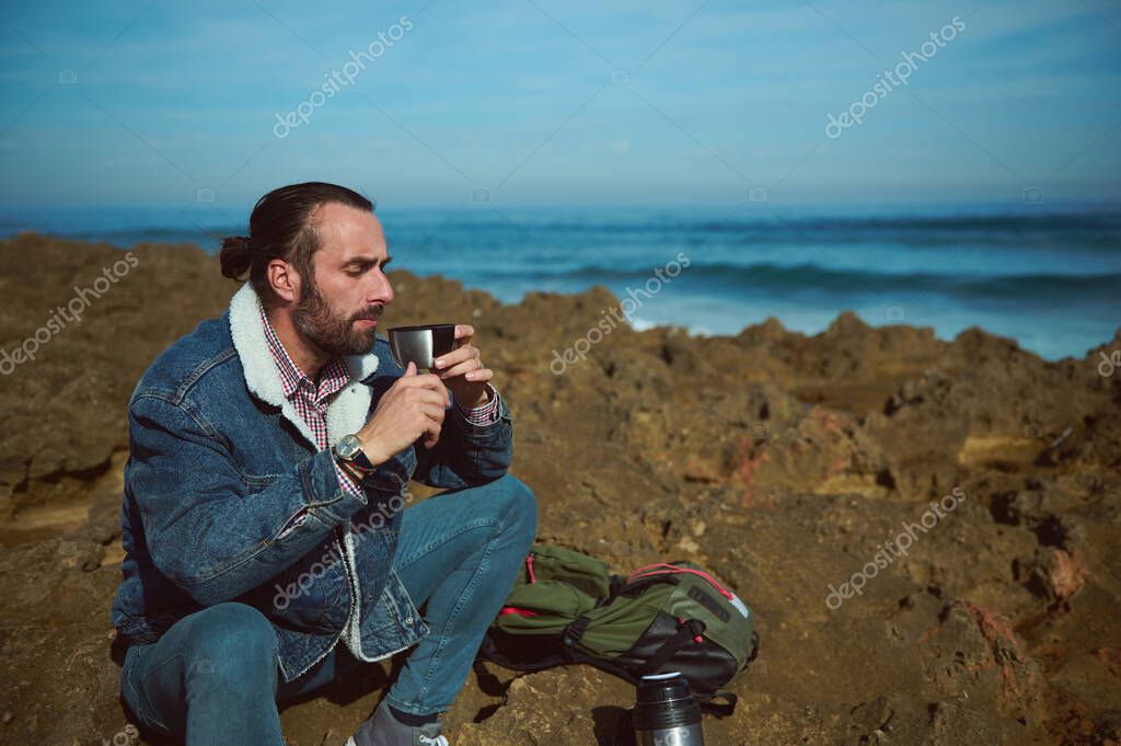 Young brutal bearded man traveling in the wild nature with thermos, have rest, enjoys his coffee break outdoor, sitting with his eyes closed on the rocky cliff by ocean, feeling connection with nature
