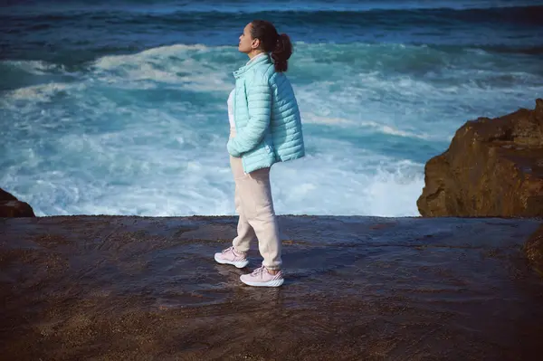 Full length portrait of a woman relaxing, enjoying connection with nature, standing back to camera on a rocky cliff, contemplating waves splashing crushing breaking on the headland. People and nature