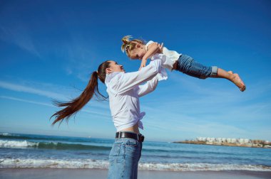 Mutlu bir anne, küçük kızını kucağında tutuyor, sevimli bir kız çocuğu uzanmış elleriyle, kusuyor ve doğada oynuyor, kumlu sahilde birlikte duruyor, dalgaların çarptığı arka planda.