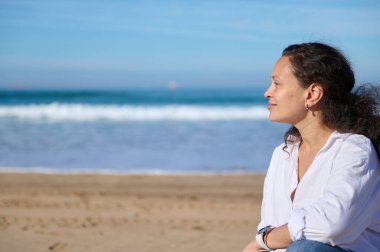 Koyu renk saçlı, rahat genç bir kadının yakından çekilmiş portresi. Uzağa bakıyor, kumlu sahilde tek başına oturuyor. Kuma vuran güzel dalgalara karşı.