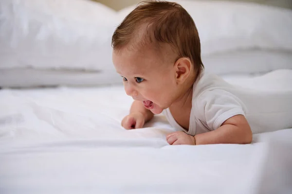 一个4 5个月大 身穿白色紧身衣 躺在肚子上笑着试图爬行的白人快乐嬉闹的小男孩的真实画像 一个快乐微笑的新生儿的画像 免版税图库图片