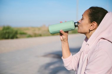 Çelik bir şişeden su içen, yoğun egzersizden sonra dışarıda dinlenen mutlu bir kadının yan portresi. Sağlıklı yaşam tarzı kavramı