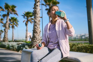 Çekici genç bayan sporcu, sıcak güneşli bir günde, şehir parkında, arka planda, taştan bir bankta oturup spor şişesinden su içerek ya da koşarak spor yaptıktan sonra dinleniyor.