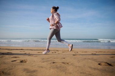 Açık havada spor yapan, koşan, güneşli bir günde kumlu bir sahilde koşan bir kadının tam boy yan portresi. Atletik kadın kıyı şeridi boyunca koşuyor. - İnsanlar. Aktif sağlıklı yaşam tarzı. Spor.