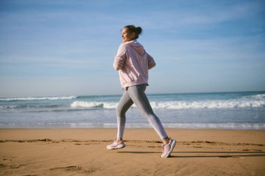 Atlantik plajı boyunca koşan orta yaşlı, etnik açıdan mutlu aktif bir sporcunun portresi. Sağlıklı bir fitness kadını dışarıda koşuyor. Spor ve sağlıklı yaşam tarzı kavramı. Tam boy kenar portresi