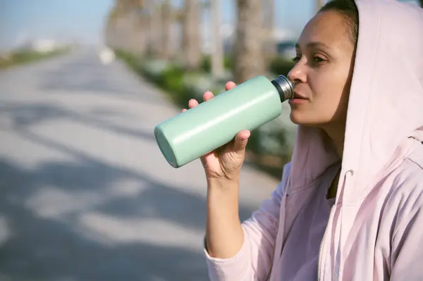 Close-up side face portrait of attractive Latina woman in light purple hoodie, holding green stainless steel bottle, drinking water to refresh her body after an outdoor workout. Copy advertising space