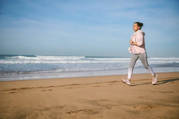 Middle aged Latin American woman jogging on the beach. People and outdoor workout. The concept of active healthy lifestyle. Full size portrait of a female athlete enjoying her morning jog on the beach